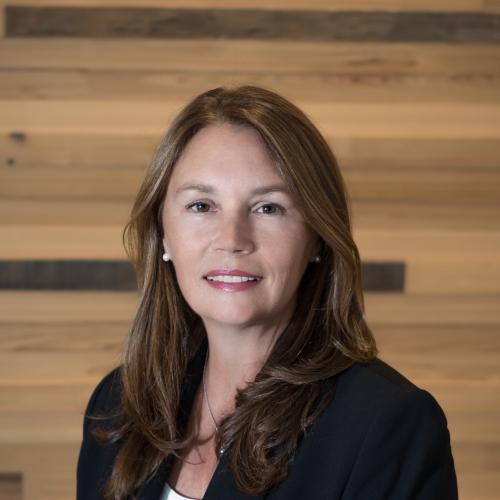 Woman smiling at camera with dark suit jacket on and wood wall behind her.