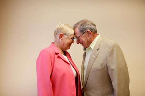 One woman in pink suit, one man in khaki suit, put foreheads together and smile.
