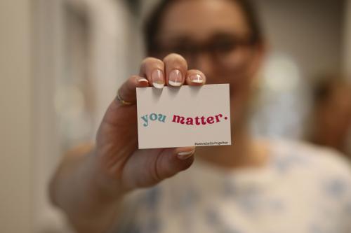 One woman with glasses holds You Matter card up close to camera with French manicure.