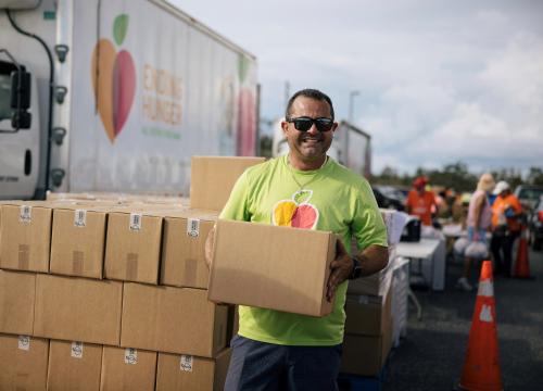 Man holds box, smiling, next to All Faiths Food Bank truck.