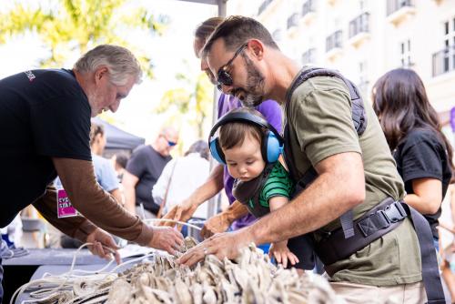 Man leans over oyster shells while baby wears headphones and watches.