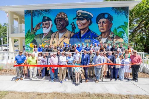 A large crowd smiles in front of a bright mural of veterans done by a local artist.