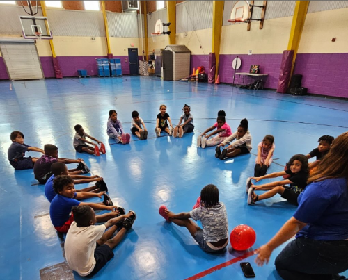 About 12 children sit on a gym floor touching their toes with a teacher instructing them.