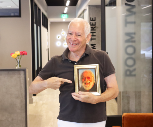 Man stands in office holding framed picture of late husband and pointing to picture.