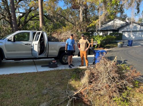 Two people stand outside by truck amidst hurricane debris.