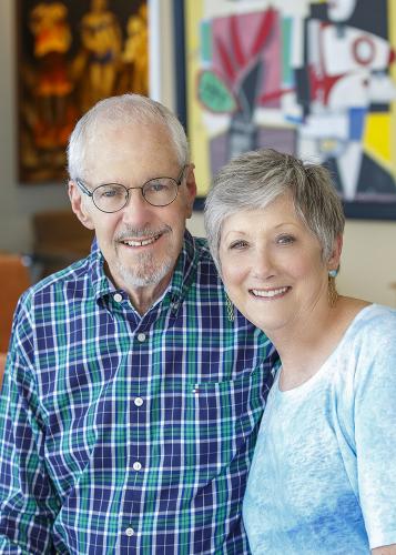 Two people, one with glasses, stand together smiling with art in background.