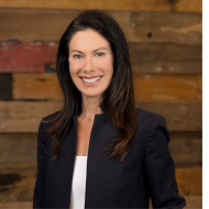 woman in a black blazer and white shirt smiles against a wood wall