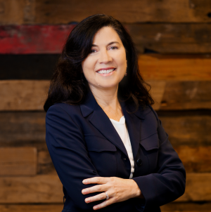 woman smiling in a black jacket and white shirt with her arms crossed against a wood wall