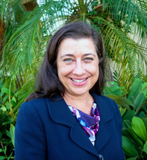 woman in a dark blue jacket with dark hair smiles at the camera in front of green bushes