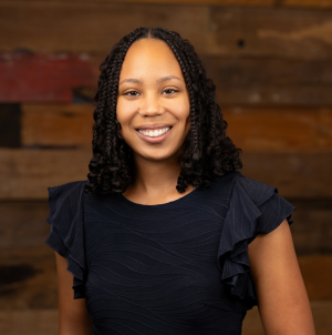 woman in a dark dress stands smiling against a wood wall
