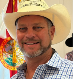 a man in a cowboy hat smiles in front of a Florida flag